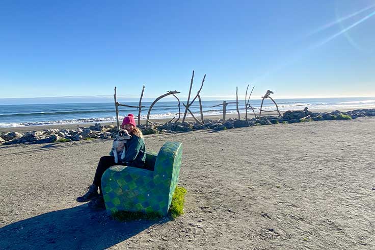 Girl sitting on armchair while holding puppy in front of Hokitika Beach stick sign